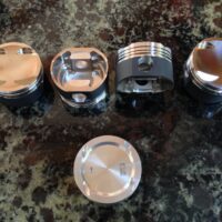 Four Forged Pistons and a piston pin displayed on a granite countertop.