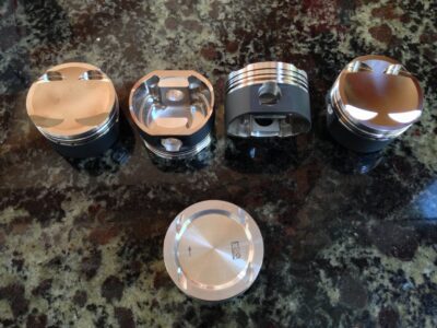 Four Forged Pistons and a piston pin displayed on a granite countertop.