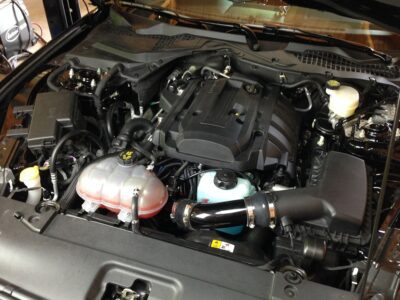 A car engine bay with various components visible, including the air intake, coolant reservoir, and engine cover.